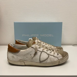 Philippe Model Prsx  - Snygga vita sneakers från Philippe Model med bruna detaljer. Skorna har en klassisk design med snörning och en bekväm passform. Perfekta för en stilren look. De är i okej skick och du kan se defekter på bild. Hör av dig för fler frågor eller funderingar.