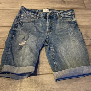 Blå jeansshorts från 157 - Snygga blå jeansshorts från 157 med slitna detaljer och uppvikta benslut. Perfekta för en avslappnad stil. De har klassisk femficksdesign och knappgylf.