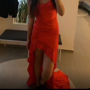 Röd långklänning med volangdetaljer - Elegant röd långklänning med volangdetaljer och asymmetrisk slits. Klänningen har en smickrande passform och är ärmlös, perfekt för festliga tillfällen.