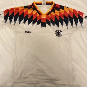 Retro Tyskland tröja  - Säljer en vit fotbollströja från Adidas med ett iögonfallande mönster i rött, orange och svart över axlarna. Tröjan har korta ärmar och en klassisk V-ringning med ränder i samma färger. Perfekt för fotbollsfans som vill sticka ut på planen!                                                                !!Byten av andra grejer funkar!!
