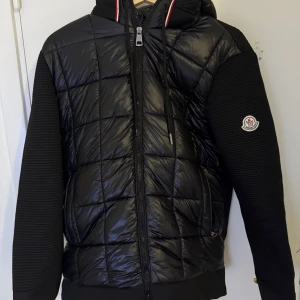 Moncler Cardigan  - Säljer en snygg svart quiltad jacka från Moncler med dragkedja och huva. Jackan har långa ärmar och ett stilrent märke på ärmen. Perfekt för kyligare dagar med sin pufferdesign och ribbade detaljer.