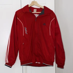 Vintage Röd vindjacka från Adidas - Säljer en röd vindjacka från Adidas med vita detaljer och dragkedja. Jackan har långa ärmar med klassiska Adidas-ränder och en broderad logga på bröstet. Perfekt för sportiga aktiviteter eller som en stilren vardagsjacka. Är från ungefär 10-20 år tillbaks!
