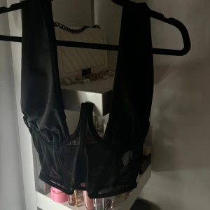 Svart korsettopp - Snygg svart korsettopp med transparenta detaljer. Perfekt för en stilren och trendig look. Toppen har breda axelband och en djup urringning.