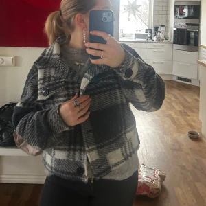 Rutig jacka i svart och grått - Säljer en snygg rutig jacka i svart och grått med stora knappar framtill. Jackan har långa ärmar, perfekt för höst och vår. Den är både stilren och bekväm, idealisk för att hålla sig varm med stil.