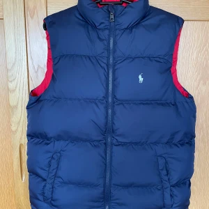 Mörkblå dunväst från Ralph Lauren - Snygg mörkblå dunväst från Ralph Lauren med röd insida och dragkedja framtill. Perfekt för kyligare dagar. Storlek Xs . Skick 9/10