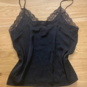Svart linne med spetsdetaljer - Snyggt svart linne med smala axelband och spetsdetaljer vid halsringningen. Perfekt för lager-på-lager eller som det är. Enkelt och stilrent plagg för många tillfällen.