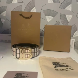 Rutig bälte från Burberry - Säljer ett stilrent bälte från Burberry med klassiskt rutigt mönster och en elegant guldspänne med logotyp. Bältet kommer i originalförpackning med tillhörande påse och presentkartong. Perfekt för att lyfta vilken outfit som helst.