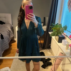 Mörkblå/grön  klänning med volangdetaljer - Snygg mörkgrön klänning med v-ringad hals och volangdetaljer. Klänningen har långa ärmar och en omlott-design. Jättefina ärmar som är lite tunnare än resten! Är lite stor på mig men verkligen så så fin. Använd typ 1 gång 
