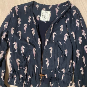 Mönstrad jacka med sjöhästar från Emma och Malena - Säljer en unik och stilren jacka från Emma och Malena med ett coolt sjöhäst-mönster. Jackan är mörkblå med dragkedja framtill och har långa ärmar. Perfekt för dig som vill sticka ut med en annorlunda stil!