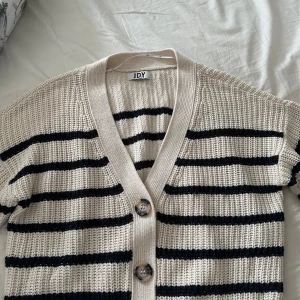 Randig kofta från JDY - Säljer en snygg randig kofta från JDY i beige och svart. Koftan har en v-ringad design med knappar framtill och är perfekt för en stilren look. Den är stickad och har långa ärmar.