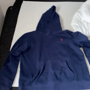 Mörkblå hoodie från Polo Ralph Lauren - Säljer en snygg mörkblå hoodie från Polo Ralph Lauren med det klassiska röda broderade logotypen på bröstet. Tröjan har en stor ficka framtill och långa ärmar. Perfekt för en avslappnad stil.