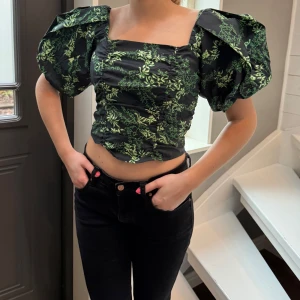 Svart och grön blus med puffärmar från Gestuz  - Snygg svart blus med grönt blommönster och stora puffärmar. Toppen har en fyrkantig urringning och en åtsittande passform som framhäver figuren