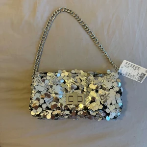 Silverfärgad axelväska med paljetter - Snygg axelväska i silver med glittrande paljetter och en kedjerem. Väskan har en stilren låsdetalj framtill och en praktisk storlek för att bära med sig det viktigaste. Perfekt för att ge en extra touch till din outfit.