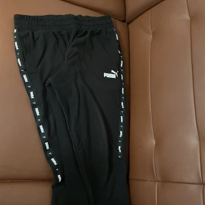 Svarta mjukisbyxor från Puma - Snygga svarta mjukisbyxor från Puma med elastisk midja och logotyp längs sidorna. Perfekta för en avslappnad stil eller träning. Byxorna har en bekväm passform och är gjorda i mjukt material.