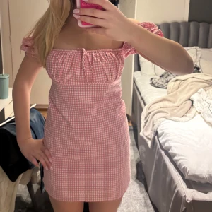 Rutig rosa klänning - Söt rosa rutig klänning med korta puffärmar och en smickrande offshoulder-design. Perfekt för sommarens varma dagar. Den har en tight passform som framhäver figuren.