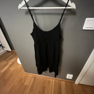 Svart klänning med smala axelband - Snygg svart klänning med smala axelband och knappar framtill. Perfekt för en stilren look. Klänningen har en enkel och elegant design som passar till många tillfällen.