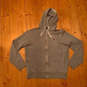 Zip up Polo Ralph Lauren - Säljer en klassisk grå hoodie från Polo Ralph Lauren med dragkedja och huva. Tröjan har en liten broderad logotyp på bröstet och ribbade muddar vid ärmslut och nederkant. Perfekt för en avslappnad stil. Liten fläck på vänster armbåge men annars felfri. Storlek M men passar S