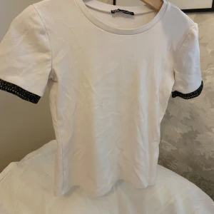 Vit t-shirt med glittriga detaljer från Zara - Säljer en vit t-shirt från Zara med korta ärmar och glittriga svarta detaljer vid ärmsluten. Jätteskönt material, Hör av dig vid frågor eller intresse 💕