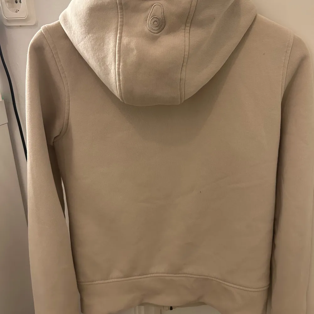 Säljer en stilren beige huvtröja från Sail Racing med dragkedja framtill och en praktisk ficka med dragkedja på bröstet. Tröjan har en bekväm huva med justerbara snören och långa ärmar. Perfekt för en avslappnad look.. Neuletakit & Villapaidat.