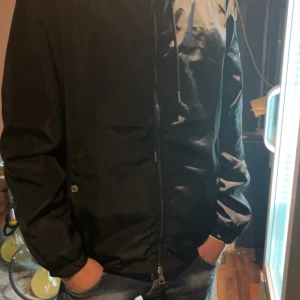 Svart jacka från Moncler - Säljer en stilren svart jacka från Moncler. Jackan har en dragkedja framtill och en hög krage med röd och vit rand. Perfekt för kyligare dagar.