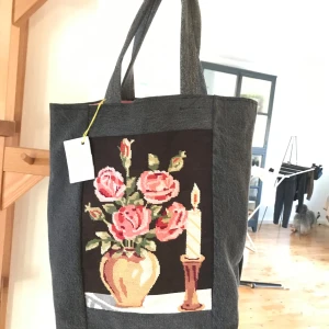 Återbrukad tygpåse jeans med broderi - Unik shoppingväska i återbrukat jeansmaterial med ett vackert broderi av rosor och ett ljus på framsidan. Perfekt för att ge din outfit en personlig touch. Väskan har en praktisk ficka på baksidan och är rymlig nog för alla dina vardagsbehov. Köpt på en julmarknad för 280 kr.