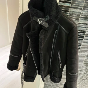 Svart jacka med pälsdetaljer - Säljer en snygg svart jacka med pälsdetaljer och dragkedjor. Den har en cool krage med spänne och är perfekt för kyliga dagar. Jackan är i mycket bra skick och passar perfekt för höst och vinter. Den har en modern och stilren design som funkar både till vardags och fest. Passar M/L storlek