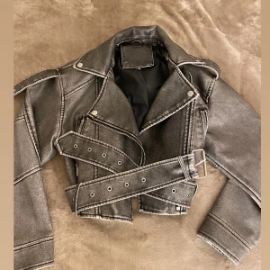 Grå skinnjacka med bälte - Säljer en cool grå skinnjacka i mycket bra skick. Den har en snygg vintage-look med dragkedja och bälte i midjan. Perfekt för höst och vår, och ger en edgy stil till vilken outfit som helst. Jackan är croppad och har långa ärmar.