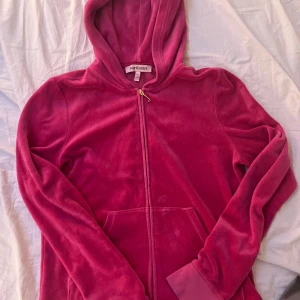 Rosa velour hoodie från Juicy Couture - Säljer en snygg rosa velour hoodie från Juicy Couture med dragkedja och huva. Den har ett stort, glittrigt JC-emblem på ryggen som ger en lyxig känsla. Perfekt för en avslappnad men stilfull look. Lite små defekter men inget som man lägger märket till.