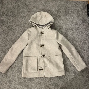 Beige duffeljacka med huva - Säljer en snygg beige duffeljacka med huva. Jackan har långa ärmar och stängs med klassiska träknappar. Perfekt för höst och vår samt vinter, och den har två stora fickor framtill. Den är i mycket bra skick och passar perfekt för en casual look.
