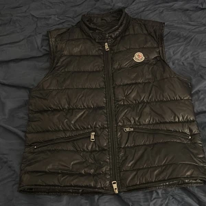 Svart dunväst från Moncler - Snygg svart dunväst från Moncler. Den har ett lagat hål på framsidan som syns i första bilden och ett olagas litet hål på ryggen som syns på sista bilden. Köpt från merch Sweden och inga kvitton finns. Skriv för fler bilder.