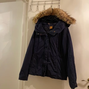 Blå parkajacka från Boss - Snygg blå parkajacka från Boss med fuskpäls på huvan. Jackan har knappar och dragkedja framtill för extra skydd mot kyla. Perfekt för kyliga dagar. Är lite förstörd har tvättat den för mycket fast går säkert o rädda om man använder tennisbollar osv 