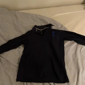 Mörkblå pikétröja från Hackett - Säljer en stilren mörkblå pikétröja från Hackett med en broderad siffra '1' i blått på bröstet. Tröjan har långa ärmar och en klassisk krage med knappar. Perfekt för en avslappnad men ändå snygg look.