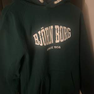 Säljer en snygg grön hoodie från Björn Borg med vit text på bröstet. Den har en klassisk känguruficka och justerbar huva med snören. Perfekt för en avslappnad stil eller träning. Passar bra till både jeans och träningsbyxor!