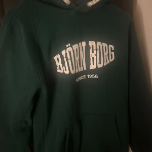 Grön hoodie från Björn Borg - Säljer en snygg grön hoodie från Björn Borg med vit text på bröstet. Den har en klassisk känguruficka och justerbar huva med snören. Perfekt för en avslappnad stil eller träning. Passar bra till både jeans och träningsbyxor!