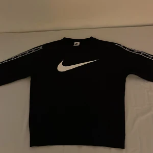 Svart tröja från Nike - Säljer en svart tröja från Nike med det klassiska Nike logan på bröstet. Tröjan har långa ärmar med vita detaljer längs ärmarna. Perfekt för en sportig look eller avslappnad stil.