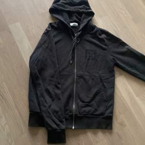 Säljer en svart hoodie från Stones med dragkedja och broderad logga på bröstet. Den har en justerbar huva och långa ärmar, perfekt för en avslappnad stil.