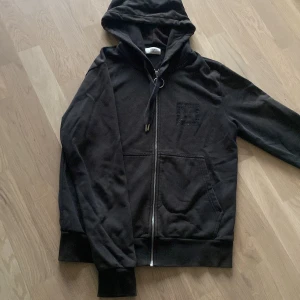 Svart hoodie från Stones - Säljer en svart hoodie från Stones med dragkedja och broderad logga på bröstet. Den har en justerbar huva och långa ärmar, perfekt för en avslappnad stil.