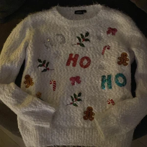 Vit jultröja från KappAhl - Mysig vit jultröja från KappAhl med glittriga bokstäver och julmotiv som pepparkakor och polkagrisar. Perfekt för att sprida julstämning! Tröjan har långa ärmar och är gjord i ett fluffigt material.