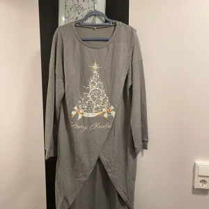 Grå långärmad klänning med julmotiv - Mysig grå lång tröja med ett julmotiv av en dekorerad julgran och texten 'Merry Christmas'. Tröjan har ett omlottmönster framtill och långa ärmar, perfekt för julfirandet.helt ny