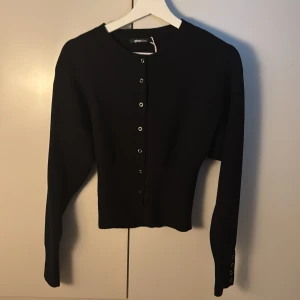 Svart kofta från Gina Tricot - Snygg svart kofta från Gina Tricot med knappar framtill. Den har långa ärmar och ribbade detaljer vid hals och ärmslut. Perfekt för en stilren look.