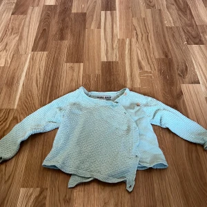 Ljusblå stickad tröja från Wai Wai - Säljer en söt ljusblå stickad tröja från Wai Wai. Tröjan har en unik omlott-design med knappar på sidan och långa ärmar. Perfekt för kyligare dagar!