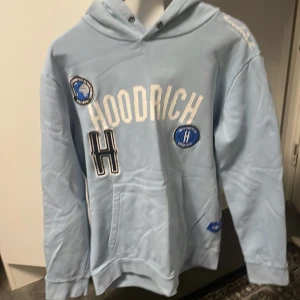 Ljusblå hoodie från Hoodrich - Säljer en snygg ljusblå hoodie från Hoodrich med coola patches och textdetaljer. Perfekt för en avslappnad stil. Den har en klassisk känguruficka och en bekväm huva. Passar perfekt till både vardag och chill.
