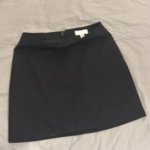 Svart kjol från H&M - Säljer denna svarta kjol från H&M i storlek 34. Kjolen har en enkel design med dragkedja bak. Den är i mycket bra skick och aldrig använd. Passar superfint till stumpbyxor. Hör av dig vid minsta fundering!