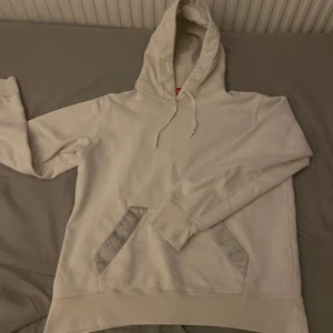 Vit supreme hoodie  - Säljer en vit hoodie av supreme i storlek medium. Den är i bra skick och har endast ett fåtal skavanker som inte syns. Riktigt snygg och jag diggar att det inte är en stor supremelogga mitt på bröstet utan det är mer diskret. Skitenkel att styla med vad som helst. Nypris ligger runt 4000kr