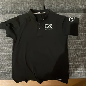 Svart pikétröja från Cutter & Buck - Säljer en stilren svart pikétröja från Cutter & Buck. Den har korta ärmar och en klassisk krage med knappar. Perfekt för både vardag och lite mer uppklädda tillfällen. Loggan syns tydligt på bröstet och ärmen. Passar bra till både jeans och chinos! Pris går alltid att förhandlas!