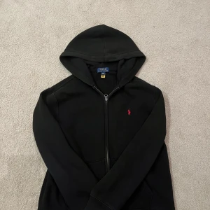 Svart hoodie från Ralph Lauren - Säljer en stilren svart hoodie från Ralph Lauren med dragkedja och den klassiska röda loggan på bröstet. Perfekt för en avslappnad look. 🖤