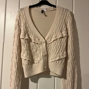 Beige kofta med spetsdetaljer - Säljer en beige kofta med v-ringning och knappar framtill. Den har långa ärmar och är dekorerad med spetsdetaljer som ger en romantisk touch. 