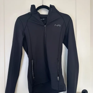 Svart tränings kofta från New Apparel - Säljer en svart kofta/jacka från New Apparel i storlek XS. Den har en dragkedja framtill och två fickor med dragkedjor. Perfekt för träning eller som en lättare jacka till vardags. Jackan är i mycket bra skick och har en stilren design med långärmade ärmar.