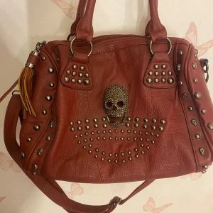Röd axelväska med dödskalle - Cool röd axelväska i läder aktigt material med en stor dödskalle och nitar som detaljer. Väskan har både handtag och en justerbar axelrem. Perfekt för en edgy look😍 Den har en dragkedja på baksidan för extra förvaring. Har även stort fack i mitten av väskan med mindre fack i sig