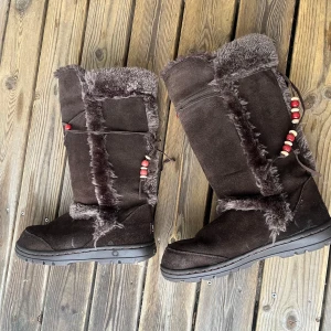 Bruna vinterstövlar från UGG - Säljer ett par mysiga bruna UGG-stövlar i mocka med fluffig pälsdetalj. De har dekorativa röda pärlor och snörning på sidan. Perfekta för kalla vinterdagar! 💖Den har mycket små defekter som man kan se på tredje bilden.